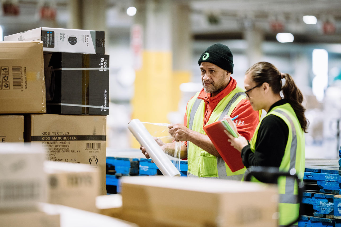 Two workers reviewing documents in a distribution center, surrounded by boxes.