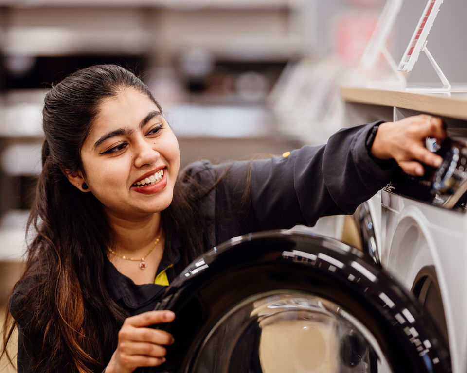 Smiling woman examines a washing machine at Noel Leeming, looking at Careers.