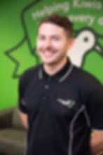 Smiling man in front of a green wall with "Helping Kiwis" text and logo